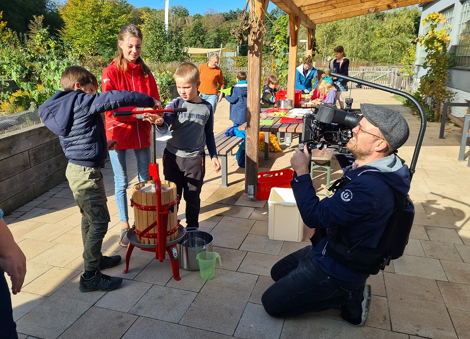 Kameramann mit Kamera filmt kniend Kinder beim Apfelpresesn mit der Handpresse
