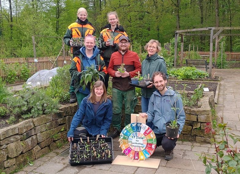 Gruppenfoto. Die Menschen halten vorgenzogene Pflänzchen und ein buntes Glücksrad in die Kamera.