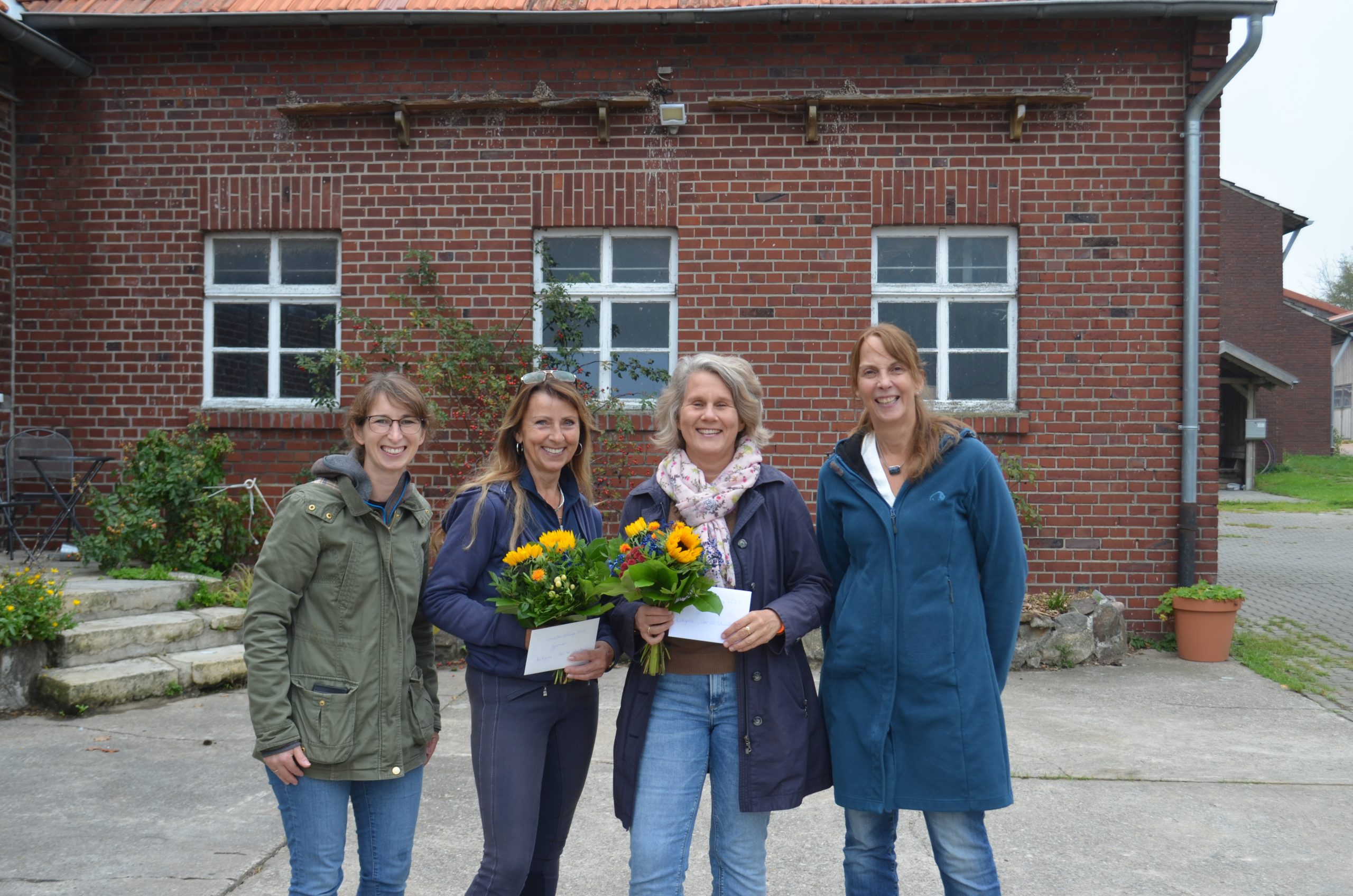 Vier Frauen. Zwei in der Mitte halten eine Urkunde und einen Blumenstrauß mit Sonneblumen.